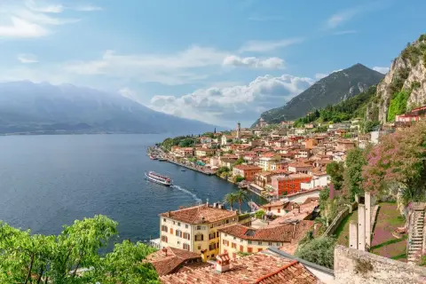Vista panoramica di una città con tetti rossi e case colorate, incastonata tra la montagna e il blu del lago di Garda. Una nave solca le acque tranquille.