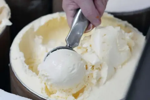 Eine Hand schöpft cremiges Vanilleeis aus einem großen Behälter. Die Eismaschine hat ganze Arbeit geleistet!