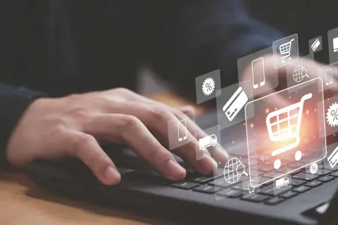 Dłoń na klawiaturze laptopa, nad nią ikony symbolizujące e-commerce: koszyk, karta płatnicza, ciężarówka, procenty, globus.