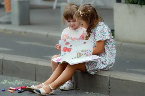 Dwie dziewczynki siedzą na krawężniku i oglądają książkę z naklejkami.