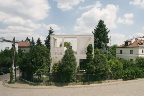 Nowoczesny dom typu kostka z białej cegły, otoczony zielenią. Widać fragmenty sąsiednich budynków i błękitne niebo z chmurami.