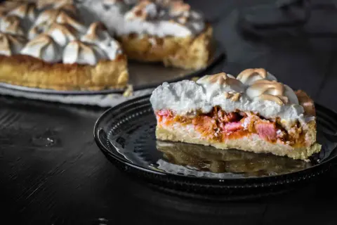 Ein Stück Rhabarberkuchen mit Baiser, das auf einem schwarzen Teller liegt. Der Kuchen hat eine goldbraune Kruste und eine saftige Rhabarberfüllung.