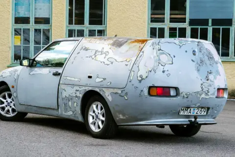 En sliten Saab 600, med rostfläckar och avskavd lack, står parkerad framför en gul byggnad med gröna fönster.