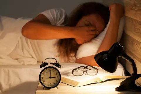 En person som gnuggar sig i ögonen i sängen, med en väckarklocka, glasögon och en bok på nattduksbordet. Känns som att vaknar flera gånger per natt.