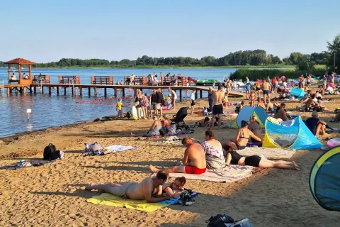 Plaża nad jeziorem powidzkim tętni życiem. Ludzie odpoczywają na piasku, kąpią się i spacerują po molo.