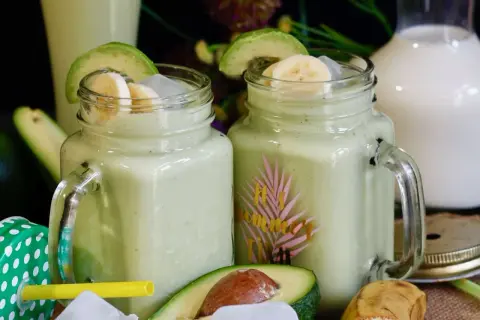 Deux verres de jus d'avocat crémeux, garnis de rondelles de banane et d'avocat, avec des fruits frais en arrière-plan.