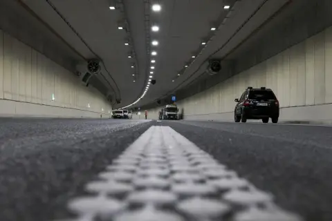 Wnętrze tunelu z widocznym ruchem pojazdów i pracami drogowymi. Widać białą linię pasów ruchu i czarny samochód. Odstęp w tunelu jest wyraźnie zaznaczony.