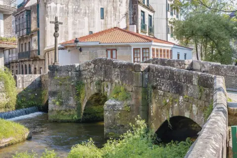 Puente de piedra con arcos sobre río, cruz de piedra y edificios históricos. Un lugar encantador que ver en Caldas de Reis.