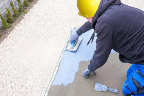 Pracownik w żółtym kasku wykonuje hydroizolację tarasu, nakładając masę izolacyjną na betonową płytę.