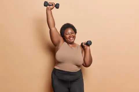 Femme souriante faisant des charges pour musculation à 70 ans, bras levés avec des haltères.
