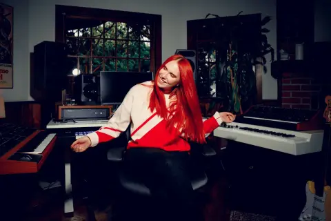 Noonie Bao, with vibrant red hair, smiles and gestures in a music studio filled with synthesizers and speakers.