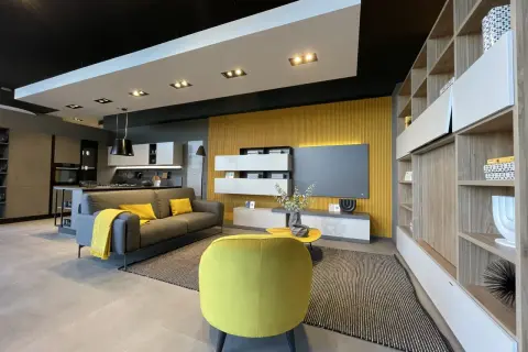 Soggiorno moderno con parete gialla, divano grigio con cuscini e plaid gialli, poltrona gialla, tavolino giallo e libreria in legno. Un esempio di colore da abbinare al giallo pareti.