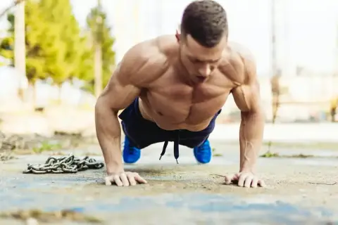 Mięśniowy mężczyzna wykonuje pompki na biceps na zewnątrz. Obok leży ciężki łańcuch.