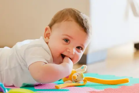 Uśmiechnięte 11-miesięczne dziecko leży na macie, gryząc paluszek i bawiąc się pluszową zabawką.