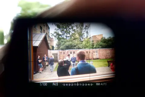 Obraz widoczny w wizjerze kamery: grupa ludzi na podwórku, jeden z nich robi zdjęcie.