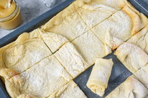Frisch gebackener Ofenpfannkuchen, in Quadrate geschnitten und mit Puderzucker bestäubt. Ein einzelnes Stück ist aufgerollt und zeigt die Füllung.
