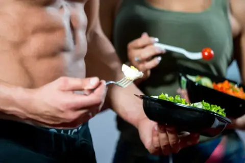 Uomo con addominali scolpiti mangia un uovo sodo, parte di una dieta ipocalorica uomo. Accanto, una donna con un'insalata.