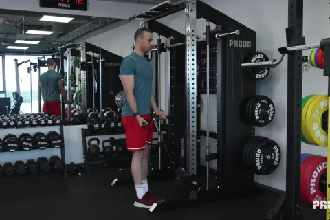 Mężczyzna wykonuje ćwiczenia na biceps przy użyciu wyciągu. W tle widoczne są sztangi, hantle i talerze obciążeniowe.