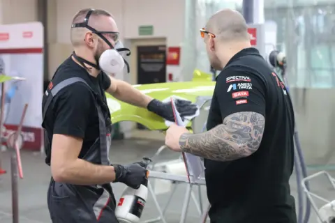 Dwóch mężczyzn w maskach i rękawiczkach pracuje nad zielonym elementem, sprawdzając, czy na podkład epoksydowy można kłaść lakier.
