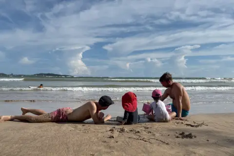 Familj njuter av en solig dag på stranden i Sri Lanka. En man ligger i sanden medan en annan leker med barnen vid havet.