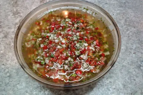 Un tazón de salsa criolla, con trozos picados de tomate, cebolla y pimiento rojo, flotando en un líquido claro.