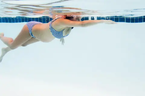En gravid kvinna i randig bikini simmar framåt i en pool. Vattnet är klart och blått.
