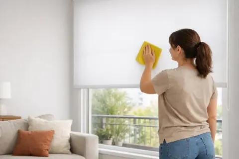 Mujer limpiando estores enrollables blancos con un paño amarillo. Consejos para como limpiar estores enrollables y mantener tu hogar impecable.