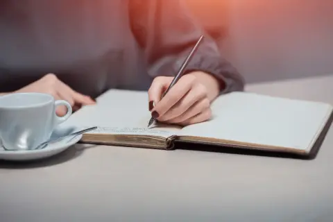 Dłoń pisze w notesie, szukając inspiracji, jak napisać intencję za zmarłego wzór. Obok filiżanka kawy.