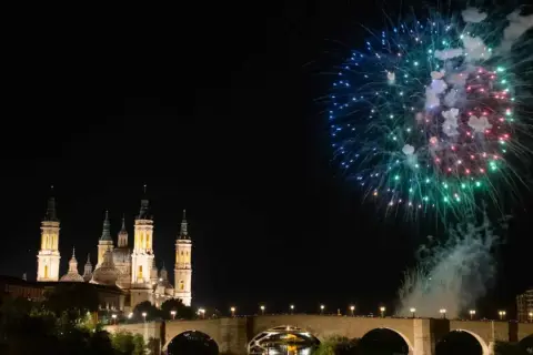 Espectaculares fuegos artificiales iluminan el cielo nocturno sobre el río Ebro, celebrando las fiestas del Pilar. La Basílica del Pilar se alza majestuosa.