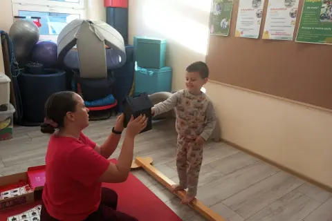 Fizjoterapeutka podaje chłopcu domek, który stoi na drewnianej belce.