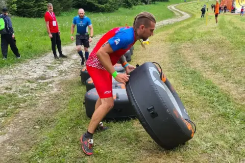 Uomo con treccia si allena al parco, spingendo un grande gonfiabile nero. Altri partecipanti corrono su un sentiero erboso.