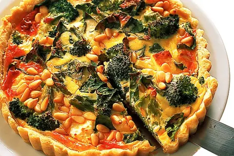 En gyllene vegetarisk paj med broccoli, spenat och pinjenötter. En bit lyfts upp med en pajspade.