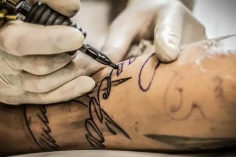 Manos enguantadas crean un diseño de letra con una máquina de tatuar. Un tatuaje con buena tinta se está formando en la piel.