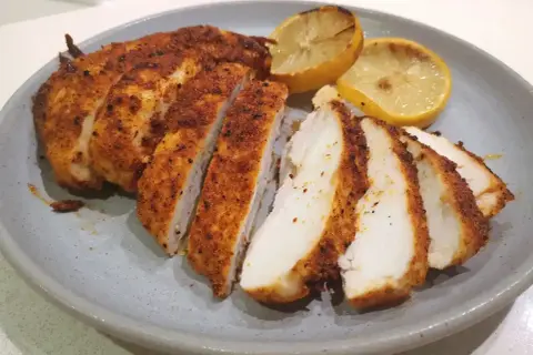 Skivad panerad kyckling med kryddig yta, serverad med grillade citronklyftor på en grå tallrik.