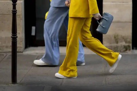 Un outfit tailleur con sneakers, giallo vivace e azzurro pastello, camminano sul marciapiede.