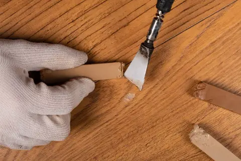 Mano guantata rimuove schegge da un parquet rovinato, mostrando rimedi fai da te per riparare il legno danneggiato.