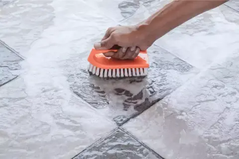 Main tenant nettoie une tache acide sur carrelage avec une brosse orange.