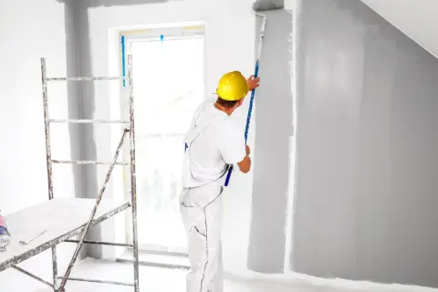 Malowanie pokoju na szaro. Pracownik w żółtym kasku wałkiem pokrywa ścianę farbą. Obok rusztowanie.