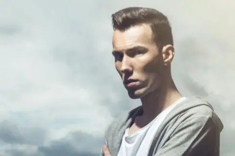 Dorian Tomasiak w szarej bluzie z kapturem i białej koszulce, z burzą chmur w tle.