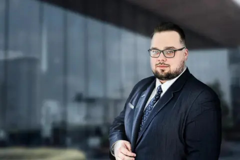 Mężczyzna w garniturze i okularach na tle nowoczesnego budynku.