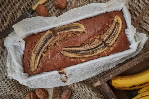 Nybakad banankaka i form, toppad med bananskivor. Valnötter och en gammal kniv ligger bredvid. Perfekt att baka med banan!