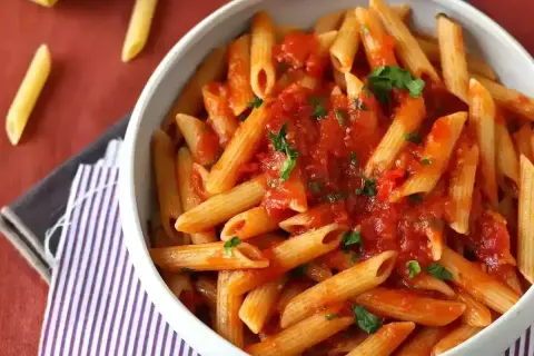 Penne all'arrabbiata w białej misce, polane pikantnym sosem pomidorowym z natką pietruszki. Obok leżą suche makarony.