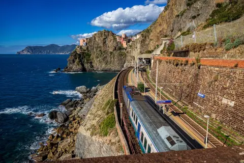 Ein Zug fährt entlang der Küste Italiens am Meer entlang. Bunte Häuser kleben an steilen Felsen.