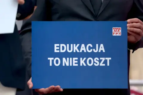 Osoba trzyma niebieską tabliczkę z napisem "EDUKACJA TO NIE KOSZT". Domaga się podwyżki dla nauczycieli 2026.
