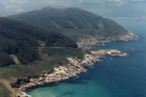 Costa rocosa y verde con el mar azul turquesa. Se ve una carretera serpenteando por la ladera. La playa Portonovo Viveiro es un paraíso natural.