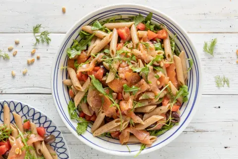 Pasta fredda con salmone affumicato, pomodorini, rucola e olive nere, guarnita con aneto fresco.