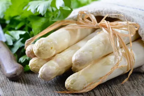 Un bouquet d'asperges blanches, prêtes à être poêlées, est noué avec une ficelle sur un tissu. Des herbes fraîches et un couteau sont visibles en arrière-plan.