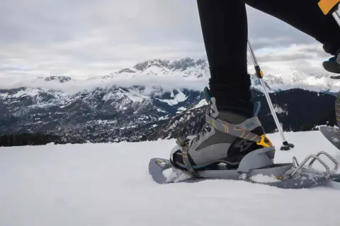 Una persona con ciaspole cammina sulla neve, con montagne innevate sullo sfondo.