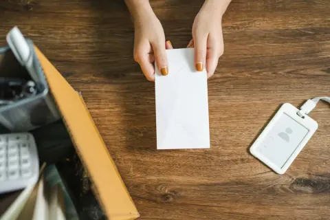 Mani che tengono una busta bianca, forse una raccomandata disdetta affitto, su un tavolo di legno con un badge e una scatola.