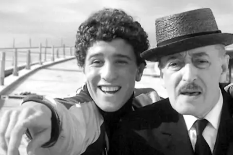 Un giovane sorridente con riccioli e un uomo anziano con cappello e baffi, un'immagine che trasmette un senso di complicità e un significato di legame generazionale.